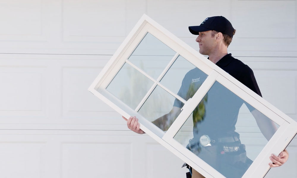 Image of man carrying a new window wearing a baseball hat in front of a garage door