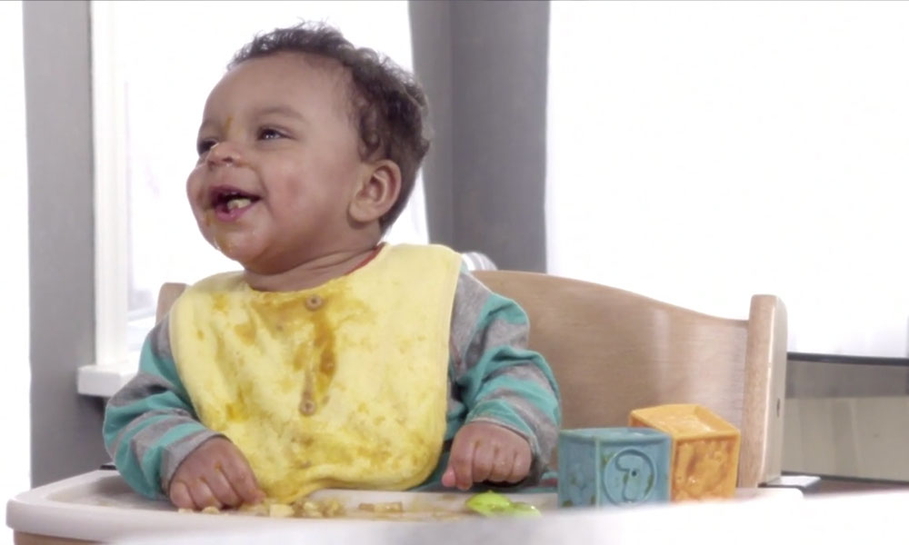 smiling baby sitting in high chair wearing a yellow messy bib with blocks and food on his tray