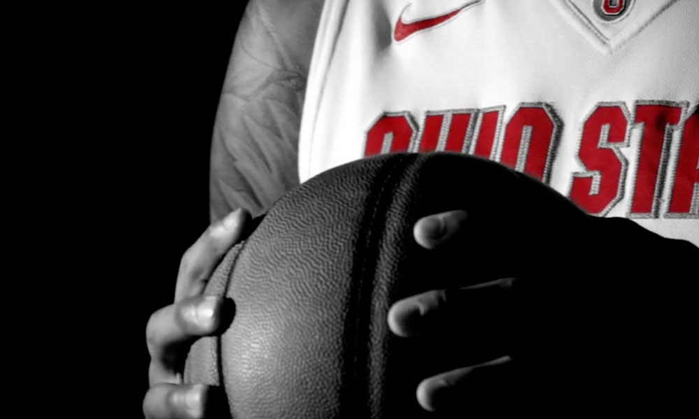 Monochrome image focusing on a person's hands holding a basketball with Ohio State University jersey in the background.