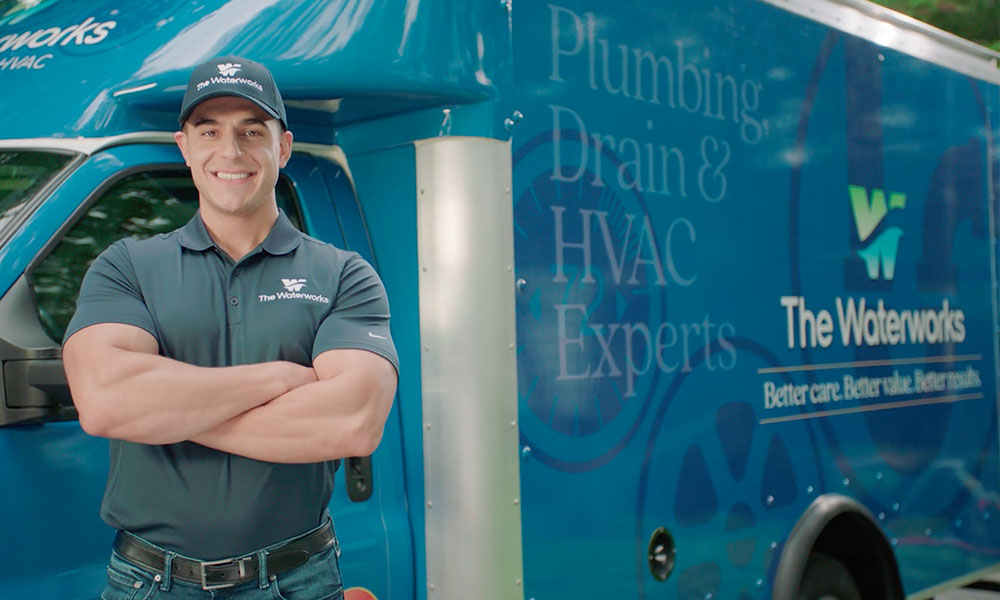 A smiling technician wearing a cap and waterworks branded t shirt standing in front of The Waterworks service truck highlighting their plumbing, drain and hvac expertise.