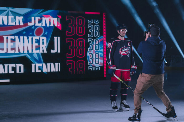 An intense behind-the-scenes shot of a hockey player being filmed, with vivid red and blue graphics displaying the player's name and number in the background.