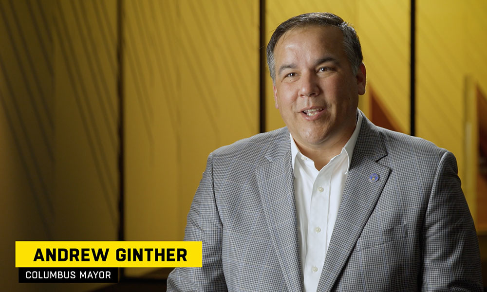 Andrew Ginther, columbus mayor, wearing a gray suit jacket being interviewed in front of a yellow back drop