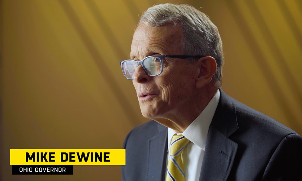 Image of older man, Ohio governer Mike Dewine, in a suit with a yellow tie and wearing blue framed glasses sitting in front of a yellow backdrop.