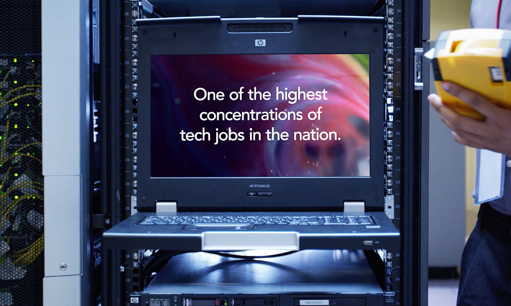 Text on a computer screen reading 'One of the highest concentrations of tech jobs in the nation' with a server rack and technician in the foreground.