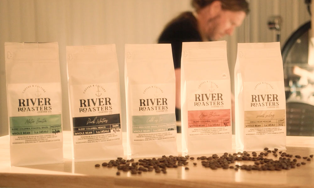 Five bags of coffee beans lined up with different colored labels indicating different roasts and beans scattered in front of each bag