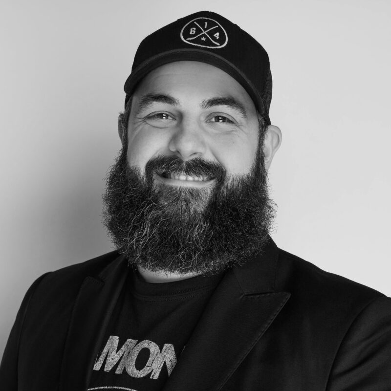 Black and white portrait of a bearded man smiling, wearing a cap and a logo t-shirt.