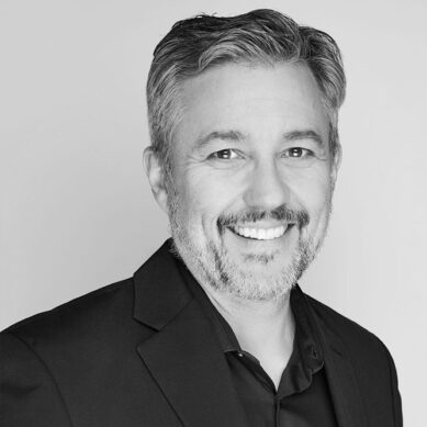 Black and white professional headshot of a smiling man with short hair and a trimmed beard and black blazer